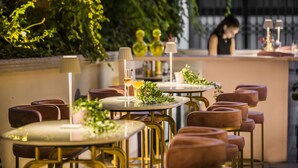 Un restaurante con mesas redondas y sillas acolchadas, algunas con lámparas. Hay plantas en macetas y una zona de bar al fondo.