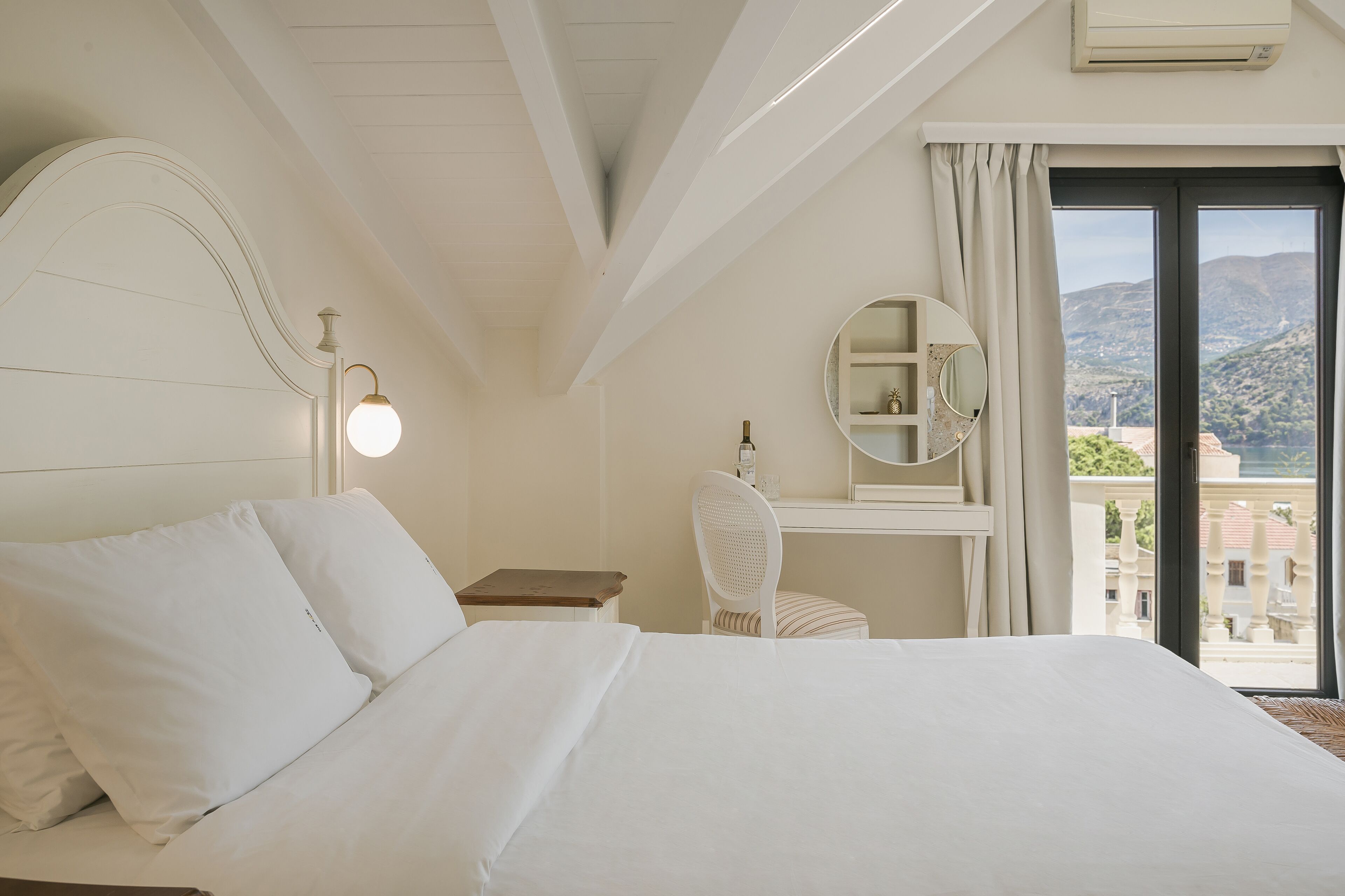 A hotel room with a large bed, a desk with a chair, a round mirror, and a view of the mountains through the window.