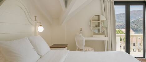 A hotel room with a large bed, a desk with a chair, a round mirror, and a view of the mountains through the window.