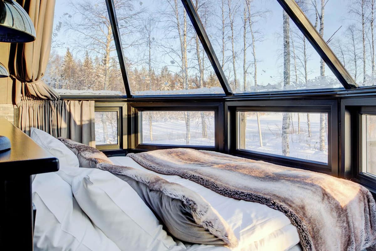 Acogedora habitación tipo cabaña con cama, lámpara y ventanas amplias que ofrecen vista a un bosque nevado.