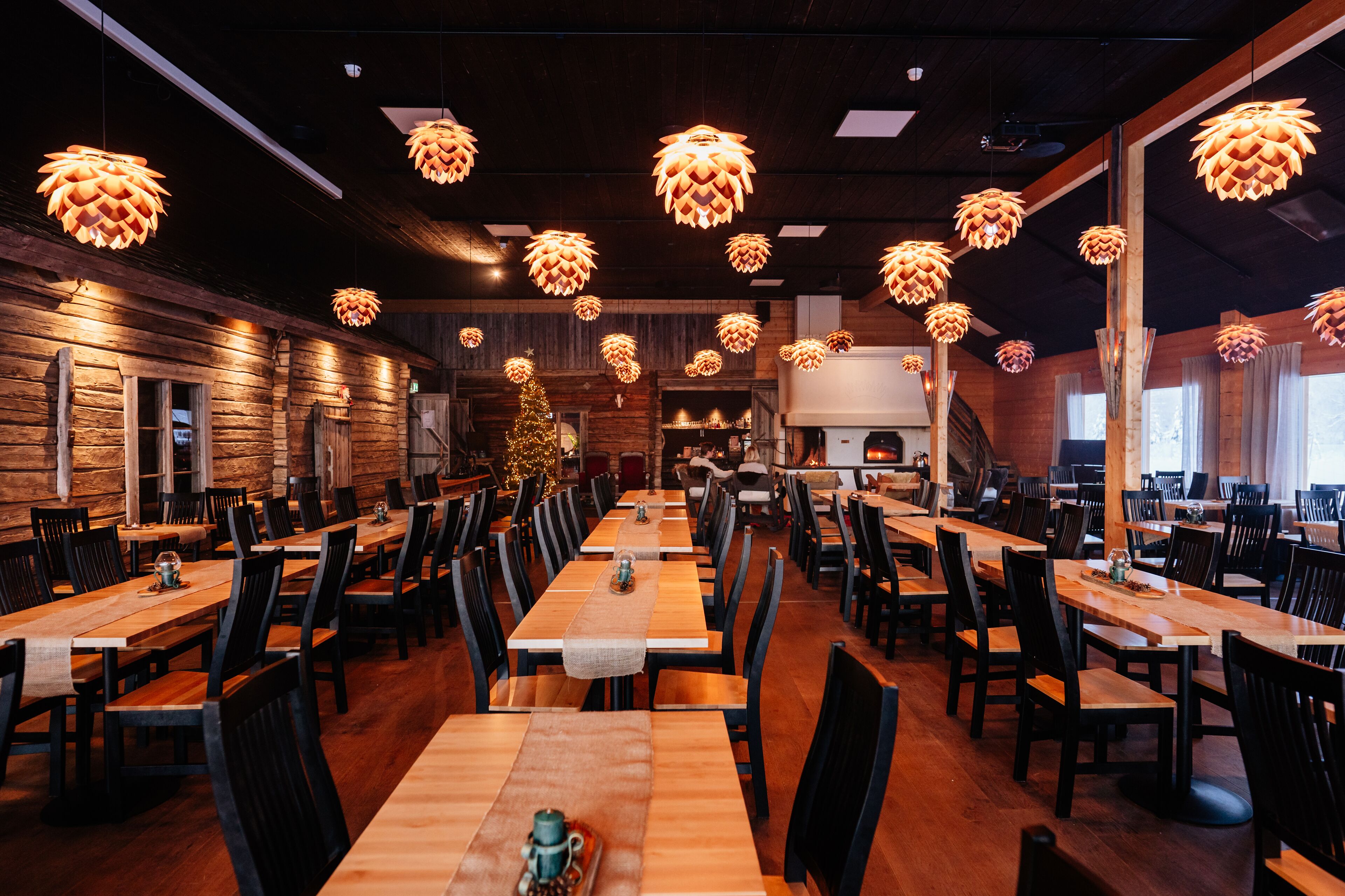 A restaurant with wooden tables and chairs, hanging pendant lights, and a Christmas tree.