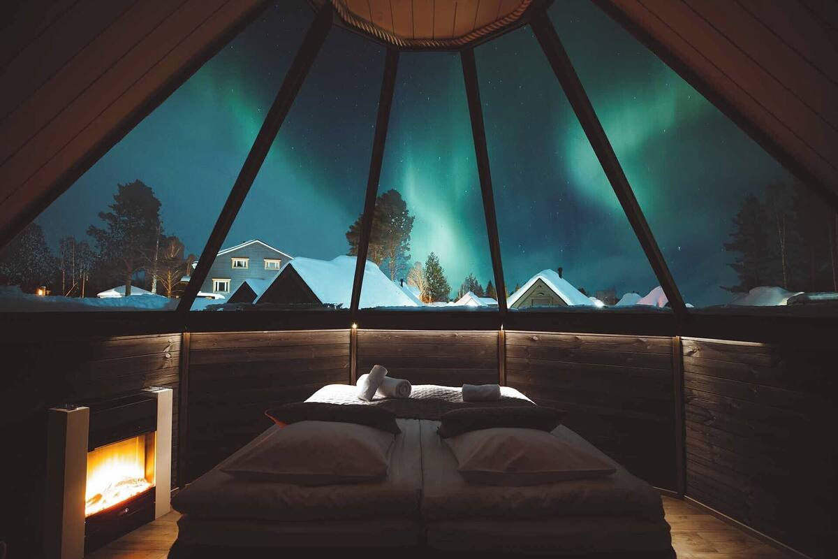 Una cabaña acogedora con cama y chimenea, que ofrece vistas de las auroras boreales sobre un paisaje nevado.