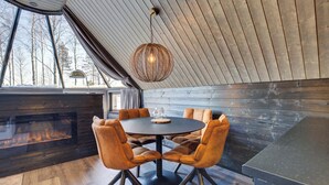 A cozy dining area with a round table and six chairs, a hanging light fixture, and a view of trees through large windows.
