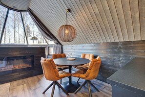 A cozy dining area with a round table and six chairs, a hanging light fixture, and a view of trees through large windows.