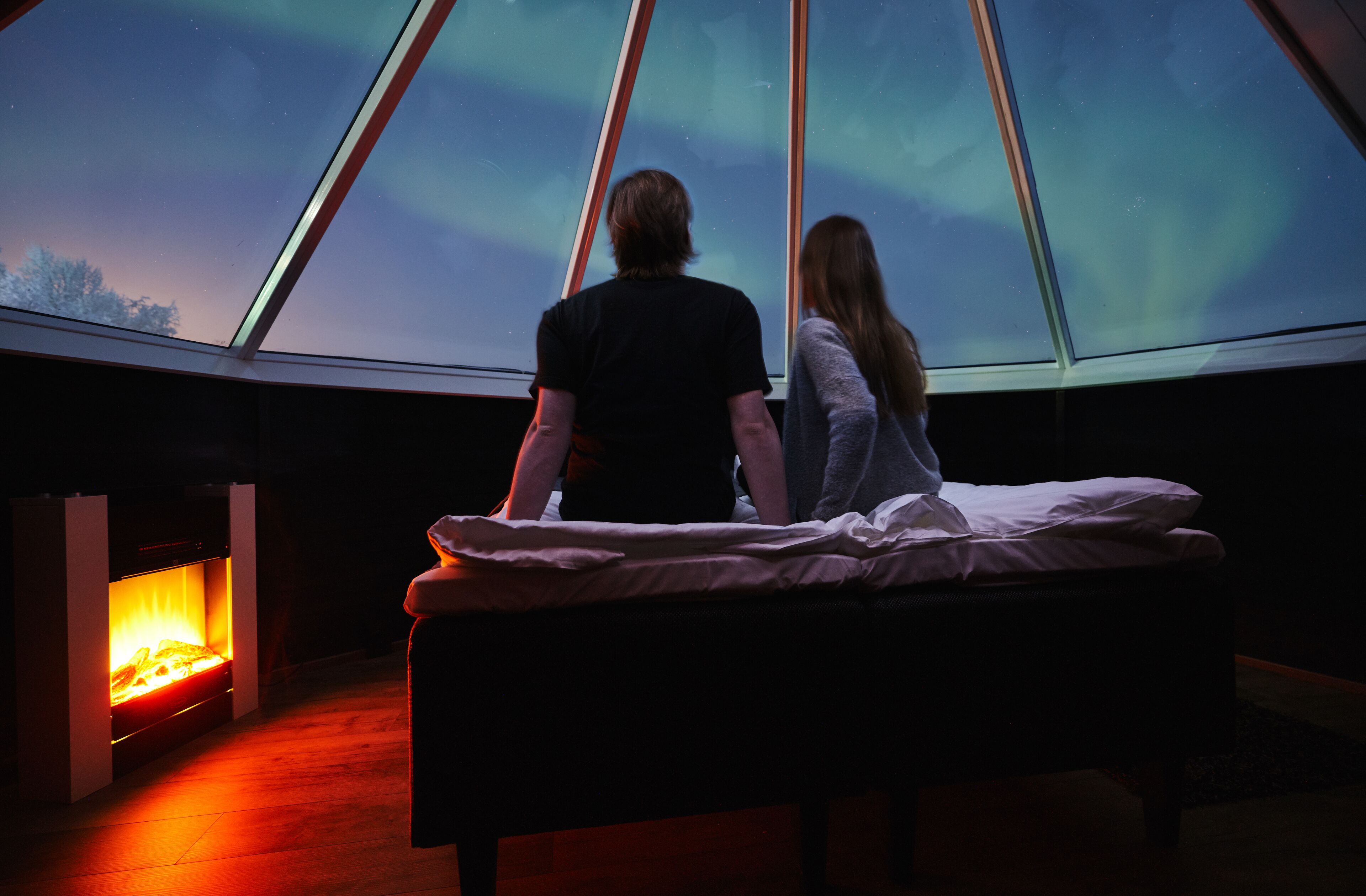 A couple in a room with a bed and a fireplace, looking out at a starry sky through a large window.