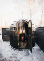 Eine Holzsauna mit Schornstein, geöffneter Tür und einer Bank im Inneren, umgeben von Schnee.