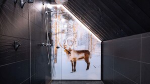 A modern bathroom with dark tiled walls, a shower, and a glass door leading to a snowy forest scene.