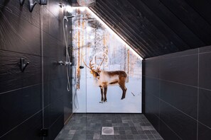 A modern bathroom with dark tiled walls, a shower, and a glass door leading to a snowy forest scene.