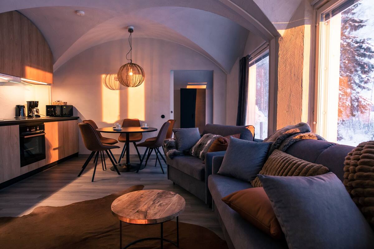 Una sala de estar moderna con un sofá, una mesa de comedor y sillas. Hay una zona de cocina con electrodomésticos y una lámpara colgante.