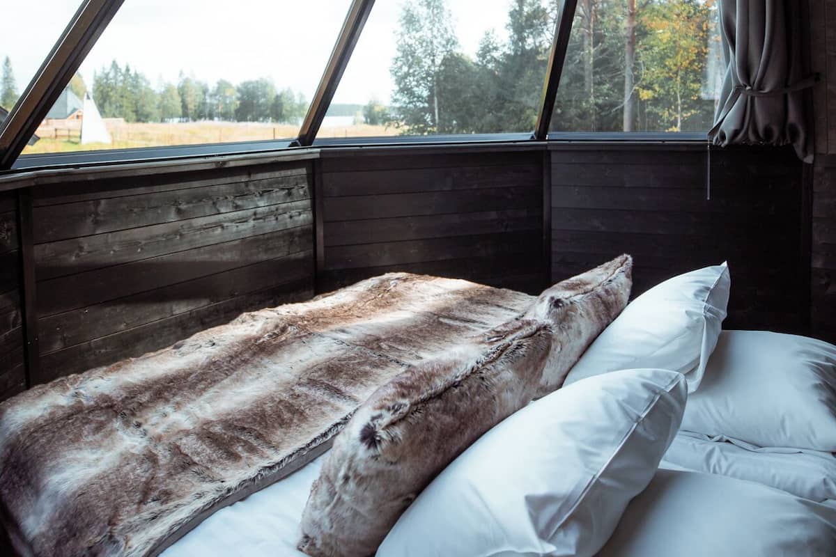 Una cama con un plaid de piel sintética y almohadas, un cabecero de madera y vistas a árboles a través de una ventana.
