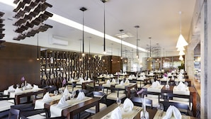 Un restaurant moderne doté de nombreuses tables, chacune recouverte d’une nappe blanche, agrémentée de serviettes et de couverts. Le plafond est orné de luminaires originaux aux formes ondulées, et les murs sont habillés de panneaux en bois.