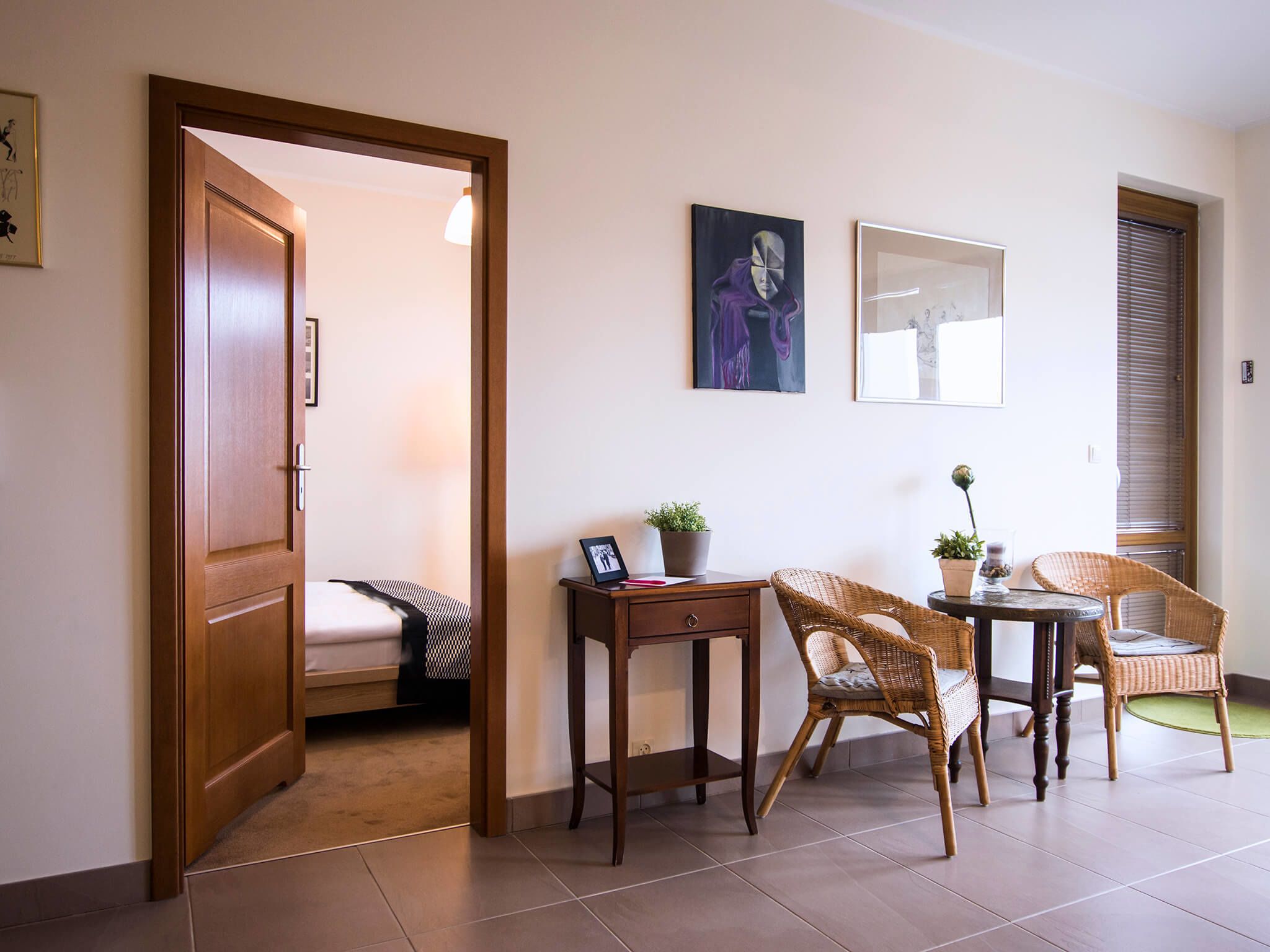 A room with a bed, a desk, two wicker chairs, a small table, and a potted plant.