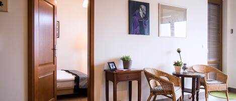 A room with a bed, a desk, two wicker chairs, a small table, and a potted plant.
