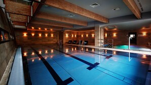 Une piscine intérieure moderne, avec des poutres en bois, un sol carrelé de bleu et un espace détente doté d’un escalier menant à un vestiaire.