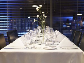 Ett långt matbord dukat med flera vinglas, en vas med vita blommor samt prydligt upplagda tallrikar och bestick.