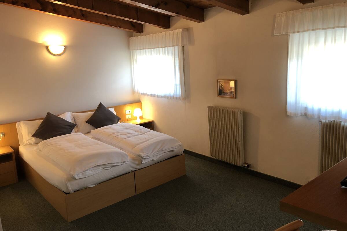 A hotel room with a wooden bed, two bedside tables with lamps, a radiator, and a small table with a chair.