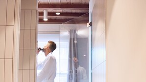 A person in a white bathrobe standing in a glass-enclosed shower area with a wooden ceiling and white tiled walls. - Maison Village (Noorbeek)