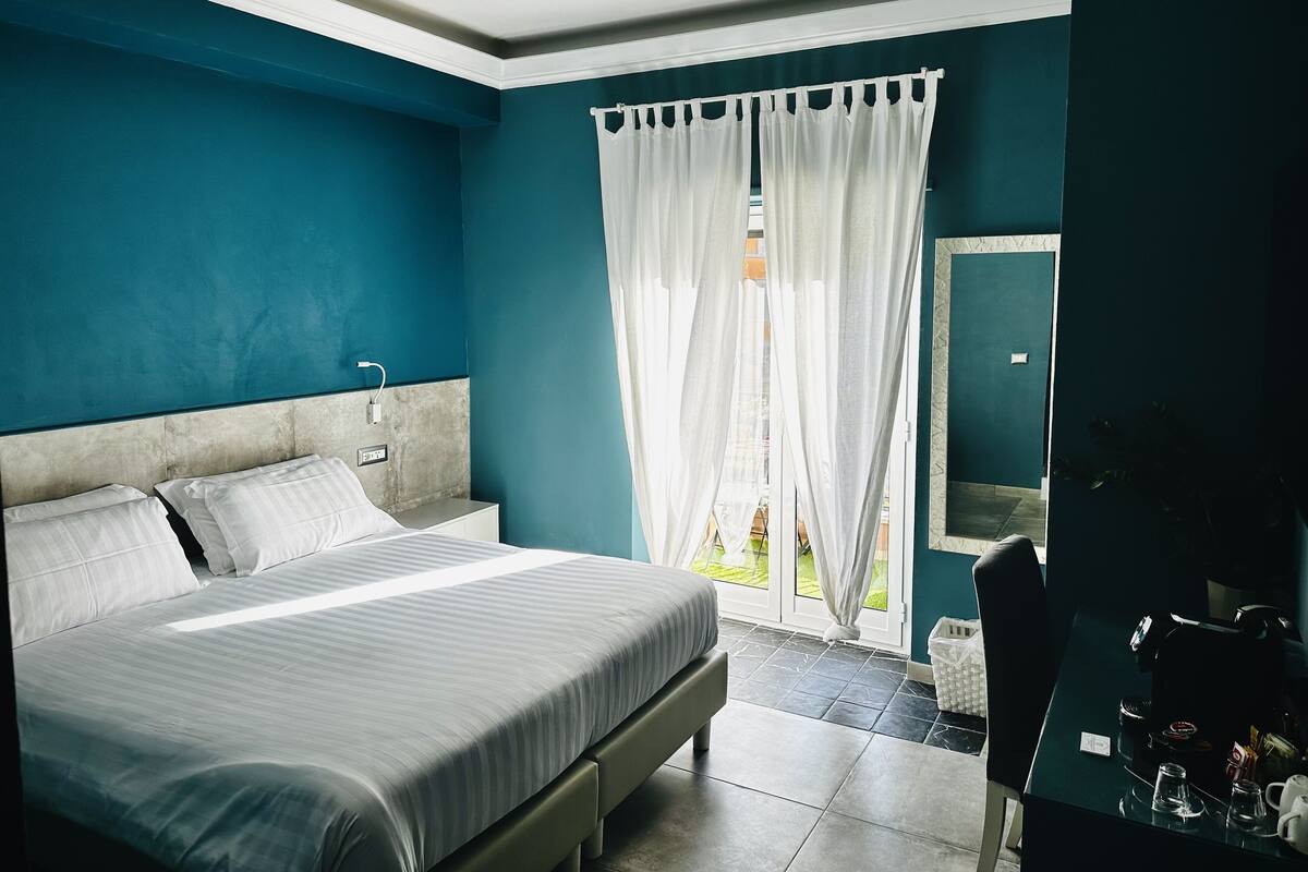 Une chambre à coucher avec un grand lit, une table de chevet, une fenêtre avec des rideaux blancs et une vue sur l’extérieur.