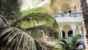 Une cour bordée de palmiers, un bâtiment blanc orné d’arcades et un balcon agrémenté d’une balustrade décorative.