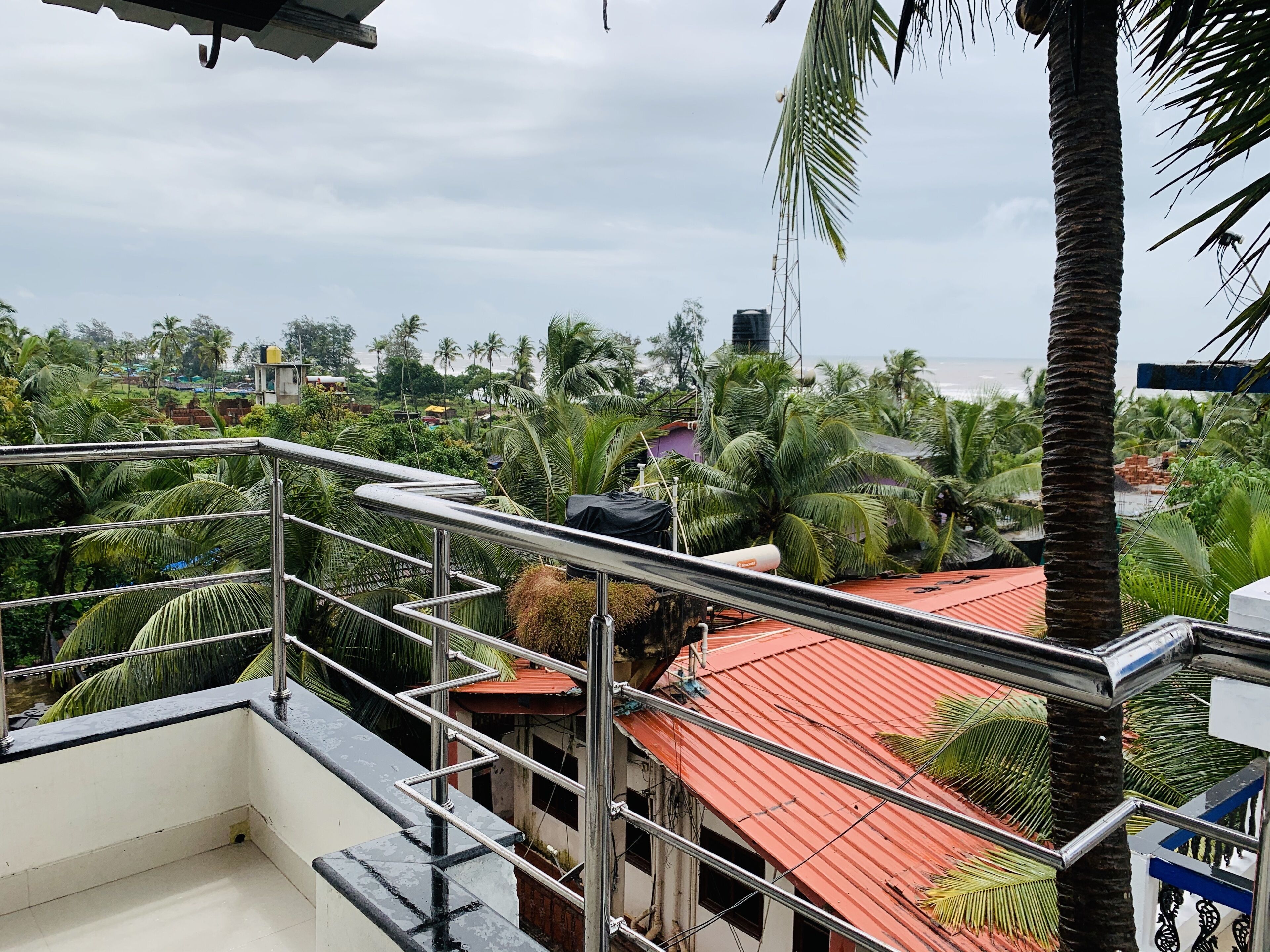 A balcony with a metal railing overlooking a residential area with palm trees and a red-roofed building.