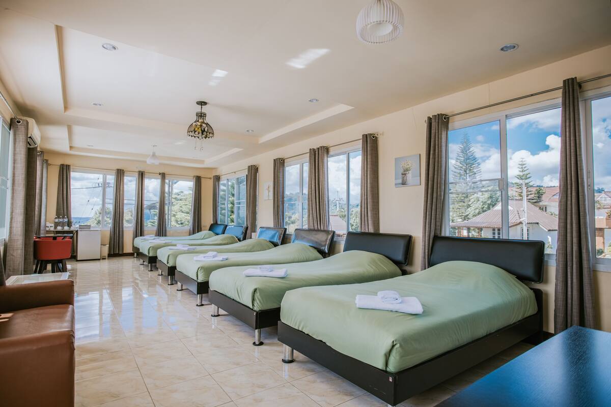 A hotel room with four single beds, a desk, a chair, and a large window with a view of trees and buildings.