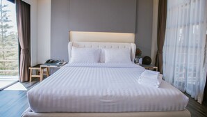 A large bed with white linens, a bedside table with a phone and a small plant, and a view of trees through the window. - Mossview Resort (Khao Kho)