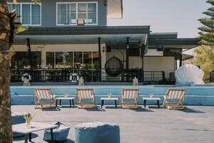 Terraza en la azotea con sillones, mesas, piscina y un edificio con amplias ventanas y un área cubierta.