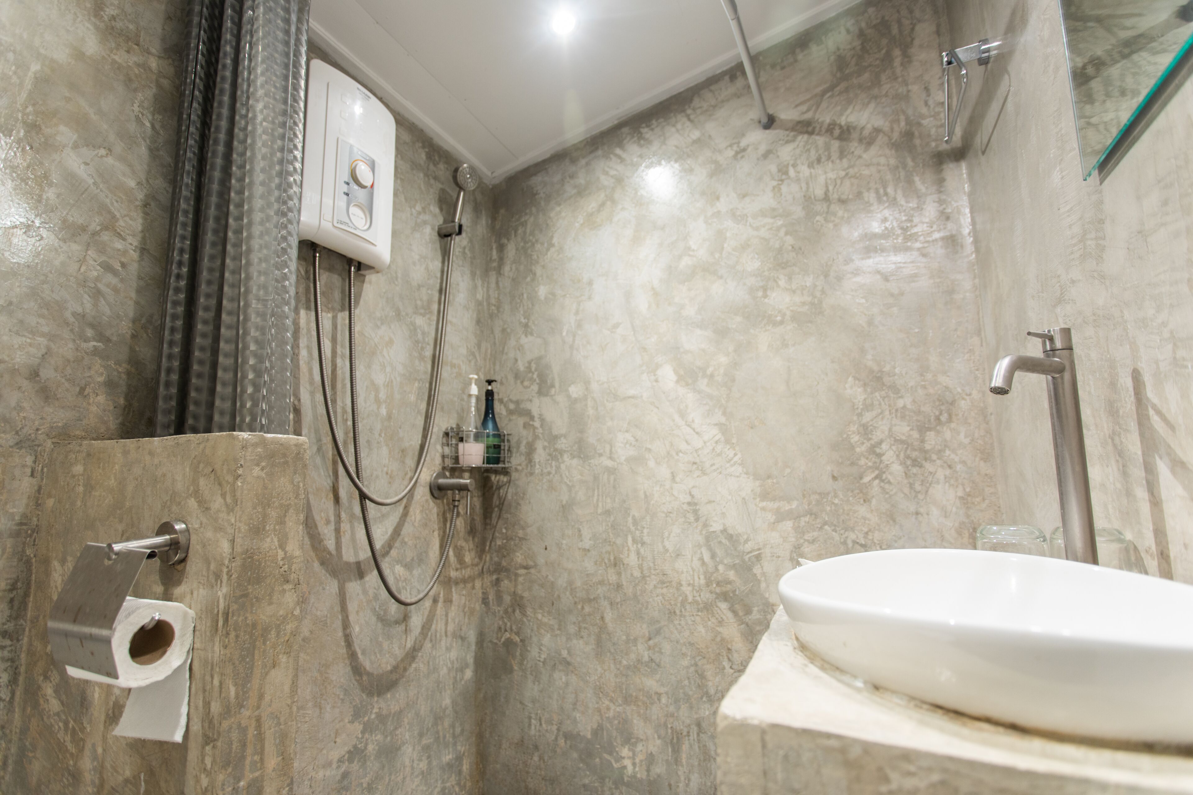 A bathroom with a marble wall, a white sink, a shower with a handheld showerhead, and a toilet paper holder.