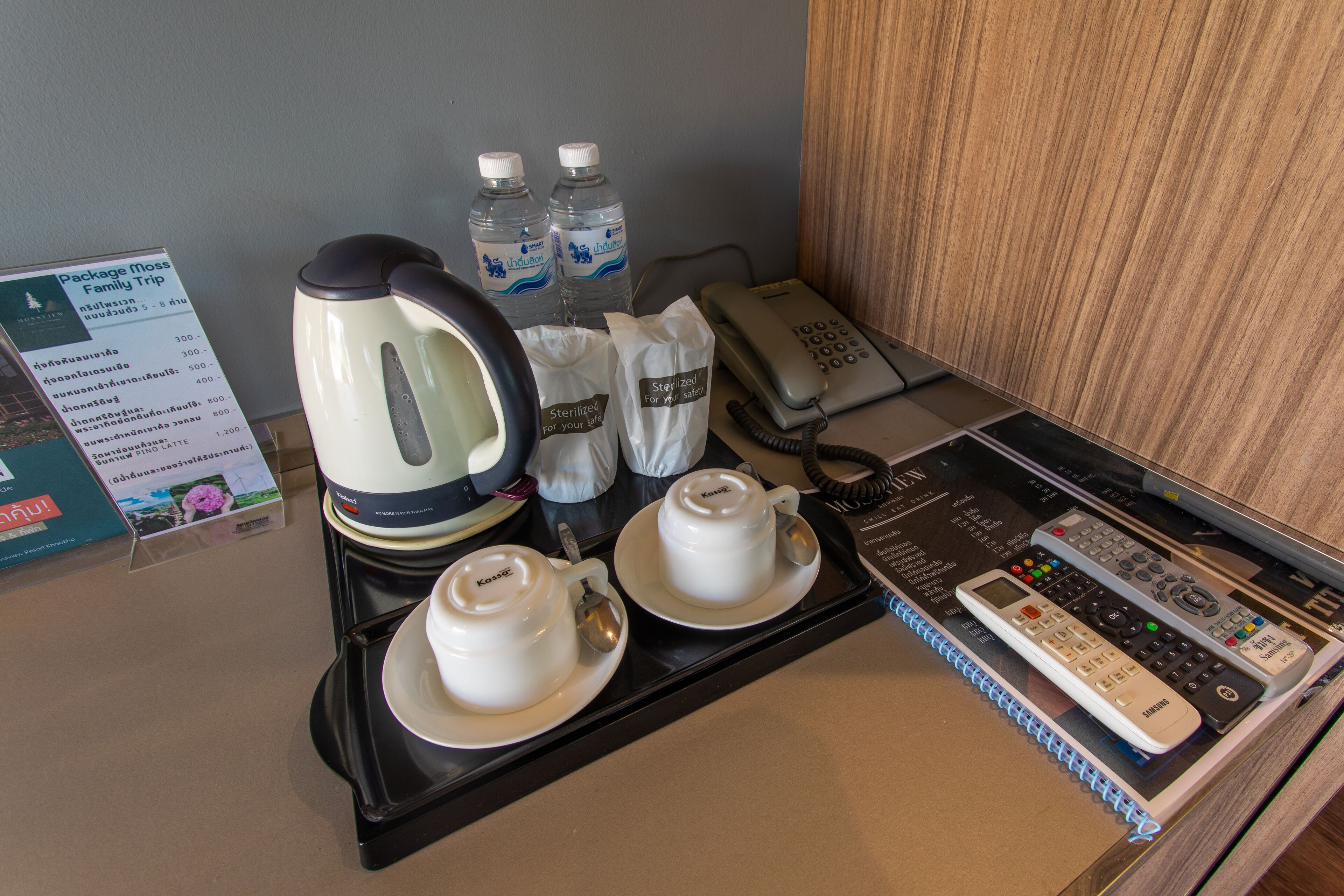 A hotel room with a coffee station, a kettle, two cups, a phone, and a remote control.