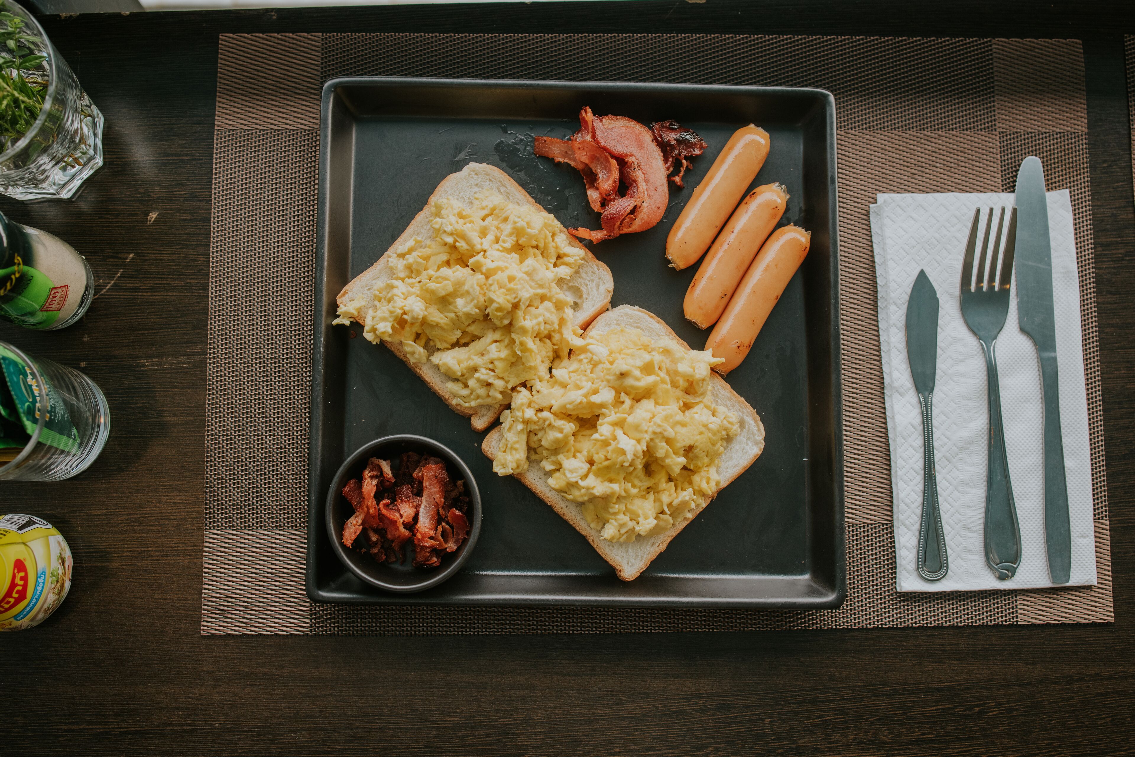 A breakfast plate with scrambled eggs, bacon, and sausages on a dark tray.