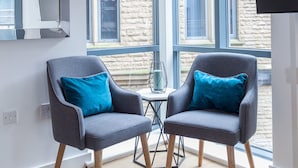 Une pièce moderne avec deux fauteuils gris, une petite table et une grande fenêtre donnant sur un bâtiment en pierre.