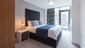 Une chambre d’hôtel moderne avec un grand lit, une table de chevet, une lampe et une vue sur la ville grâce à une grande fenêtre.