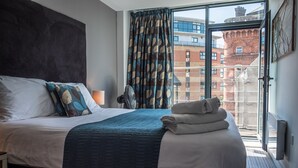Une chambre d’hôtel moderne avec un grand lit, une vue sur un immeuble en briques par la fenêtre et un balcon doté d’une rambarde en métal.
