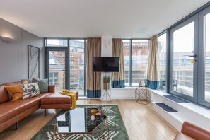A modern living room with a leather sofa, a glass coffee table, and a large window offering a city view. - Kspace Serviced Apartments Leeds (Leeds)