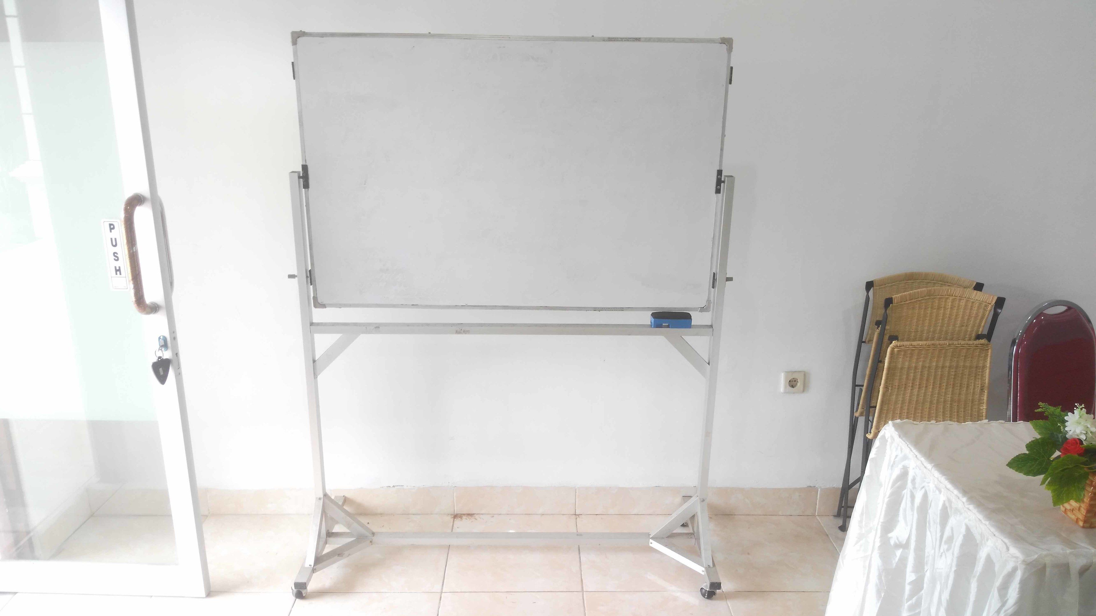 Un atril para pizarra en una habitación con puerta de vidrio, una silla de mimbre y una mesa con mantel blanco y un jarrón con flores.