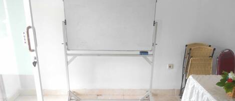 Ein Whiteboard steht in einem Raum mit Glastür, einem Korbstuhl und einem Tisch mit weißem Tuch und einer Blumenvase.