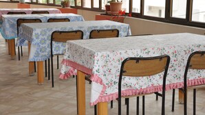 Una hilera de mesas con manteles florales y bordes festoneados, sillas de madera y plantas en macetas sobre un piso de baldosas, con grandes ventanales al fondo.