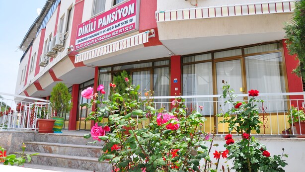 Un edificio con un letrero que dice "DİKİLİ PANSİYON" e información de contacto. Hay escalones que llevan a la entrada, y macetas con flores en la parte delantera.