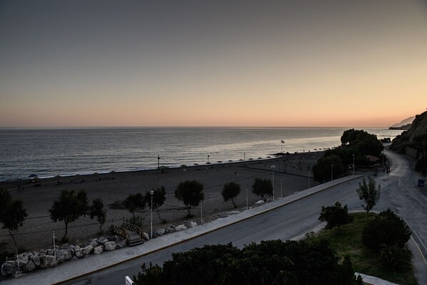A coastal road with a beach on the right, a pedestrian path, and a rocky area with trees. - Ariadni Hotel by Estia (Viannos)