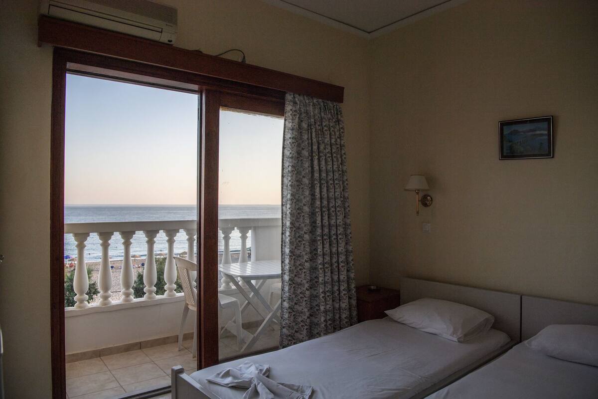 Ein Hotelzimmer mit Bett, Nachttisch, Lampe, Fenster mit Blick auf den Strand und einem Balkon mit Tisch und Stühlen.