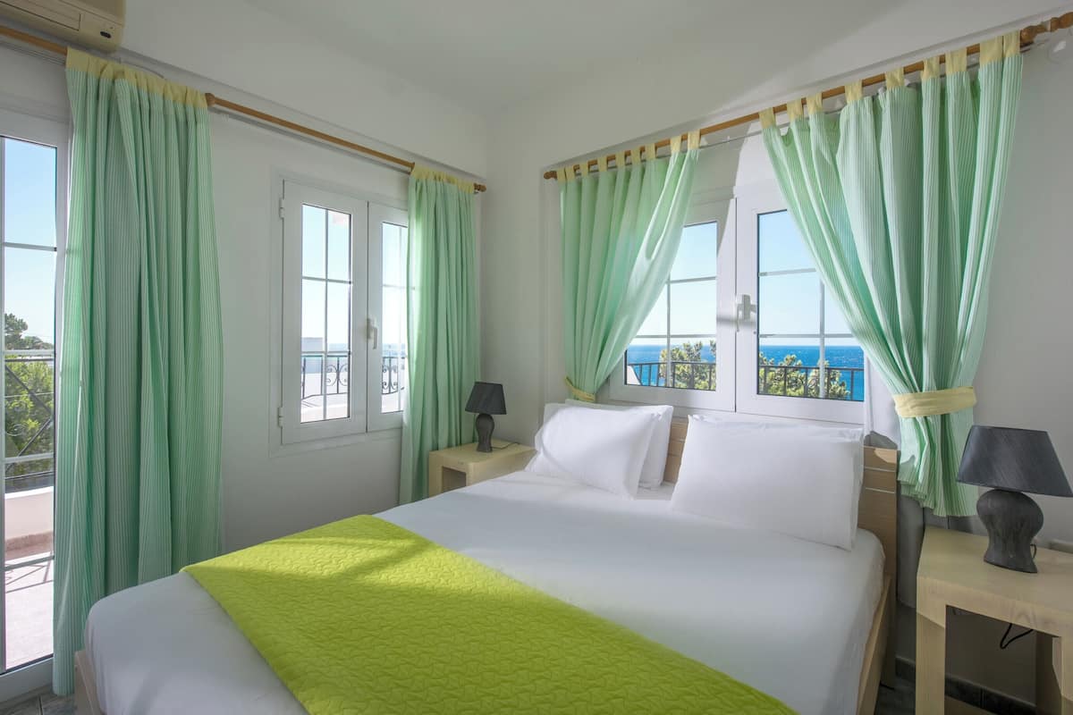 Een hotelkamer met een bed, twee nachtkastjes met lampen, een raam met gordijnen en uitzicht op zee.