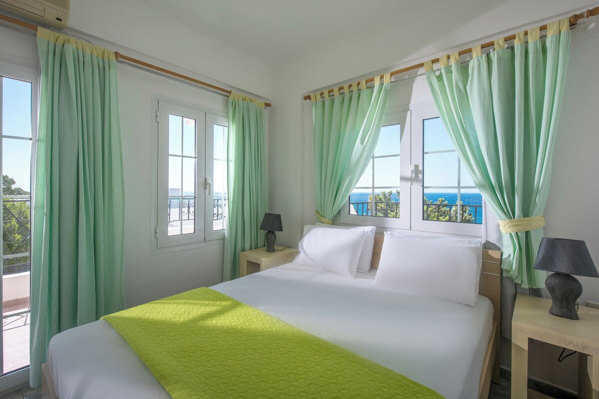 Een hotelkamer met een bed, twee nachtkastjes met lampen, een raam met gordijnen en uitzicht op zee.
