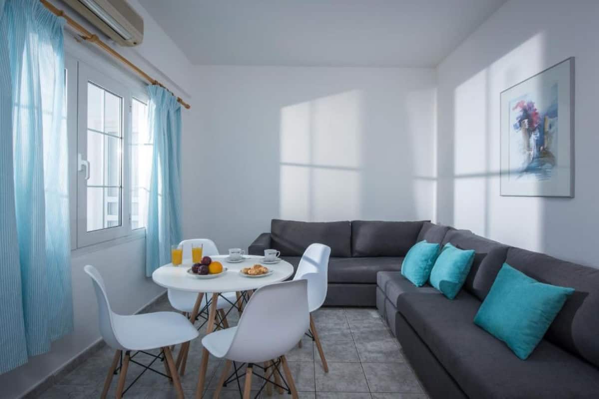Een moderne woonkamer met een grijze bank, witte stoelen, een ronde tafel met ontbijtartikelen en een raam met gordijnen.