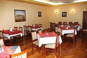 Un espace repas comprenant plusieurs tables, chacune recouverte d’une nappe blanche et agrémentée de serviettes orange, situé dans une pièce au sol en bois et aux murs de couleur beige.