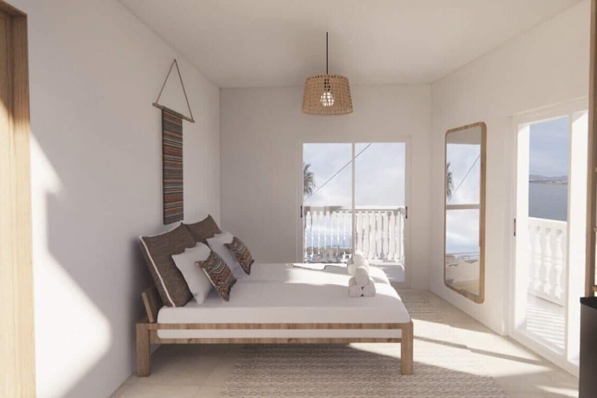 A bedroom with a large bed, a hanging light fixture, a view of the sea through a window, and a balcony with a railing.