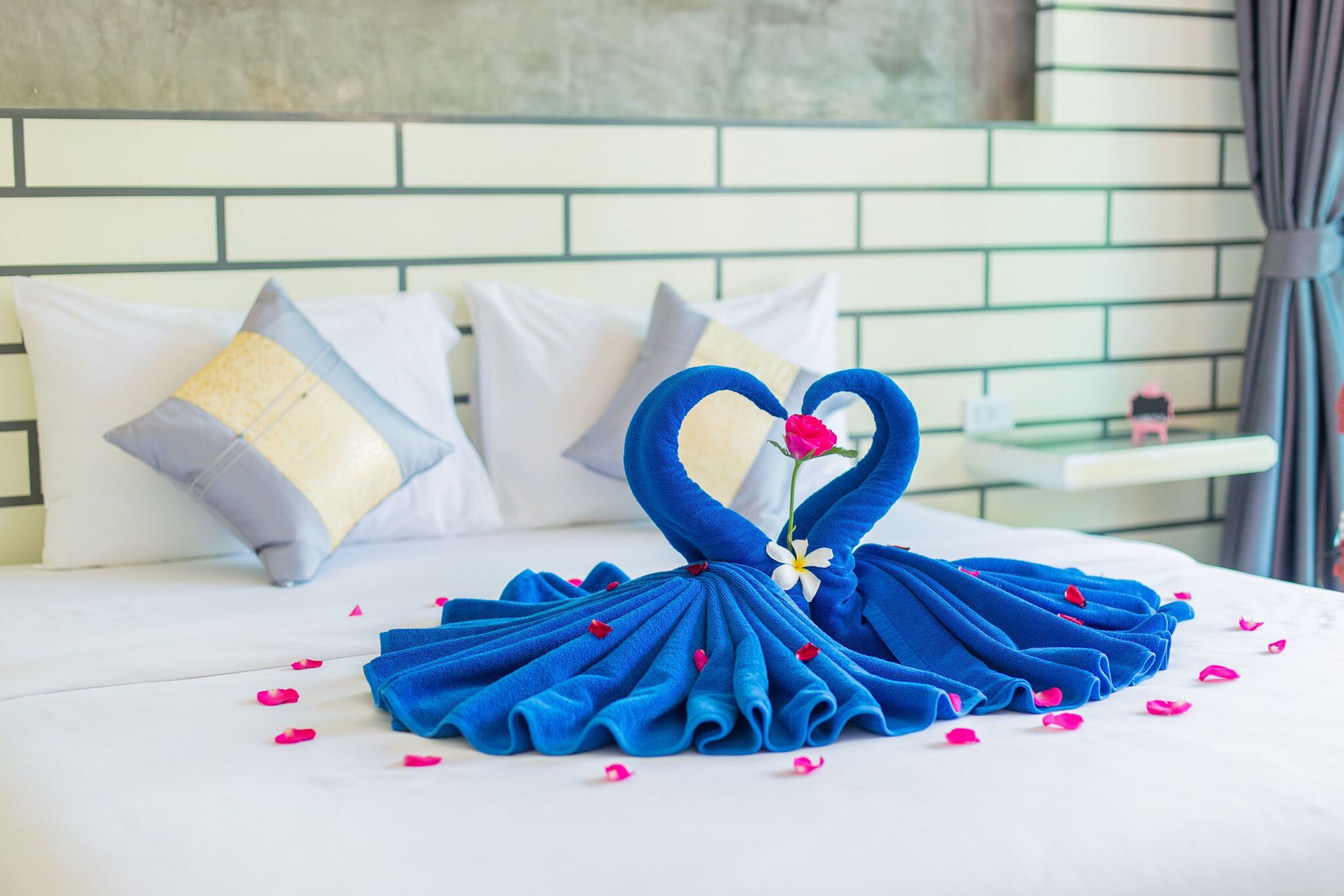 Un lit agrémenté d’une décoration en forme de cœur réalisée avec des serviettes et des pétales de rose, avec une literie blanche et une tête de lit à motifs de briques.
