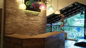 Una zona de recepció amb un taulell de fusta corbat, llums penjants, un ventilador de sostre i un mural a la paret de maó.