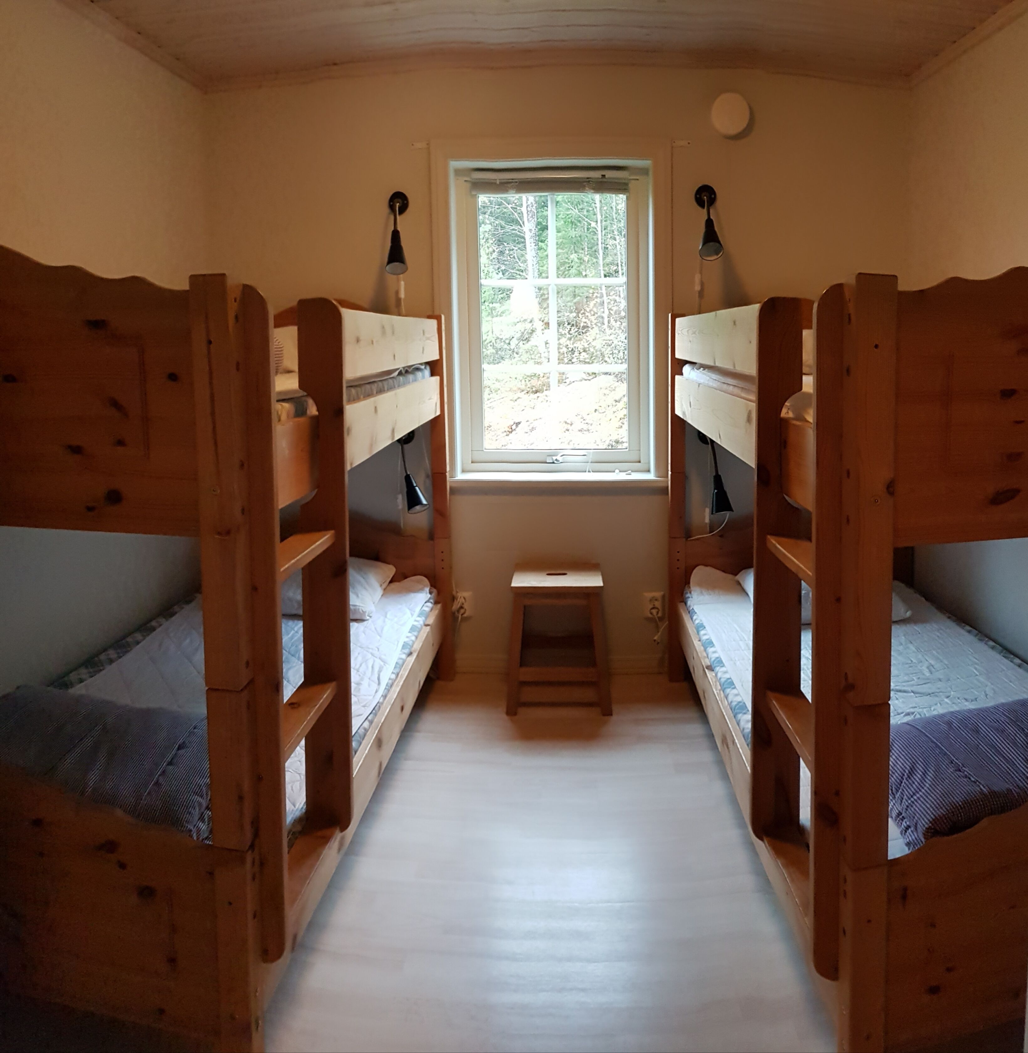 A room with two wooden bunk beds, a small table, and a window with a view of trees.