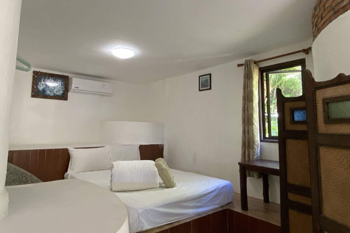A hotel room with a bed, a bedside table, a window with curtains, an air conditioning unit, and a picture on the wall.
