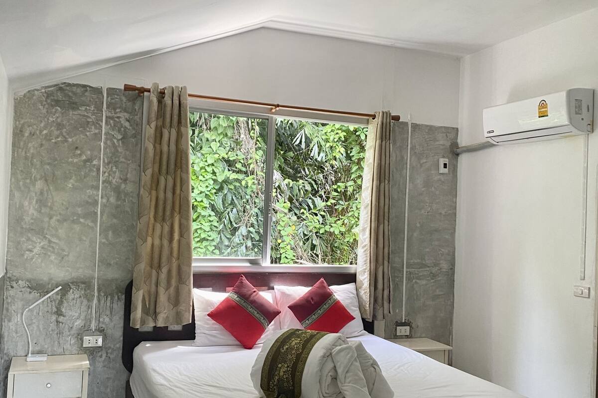 A bedroom with a bed, a nightstand, an air conditioning unit, and a window with curtains.